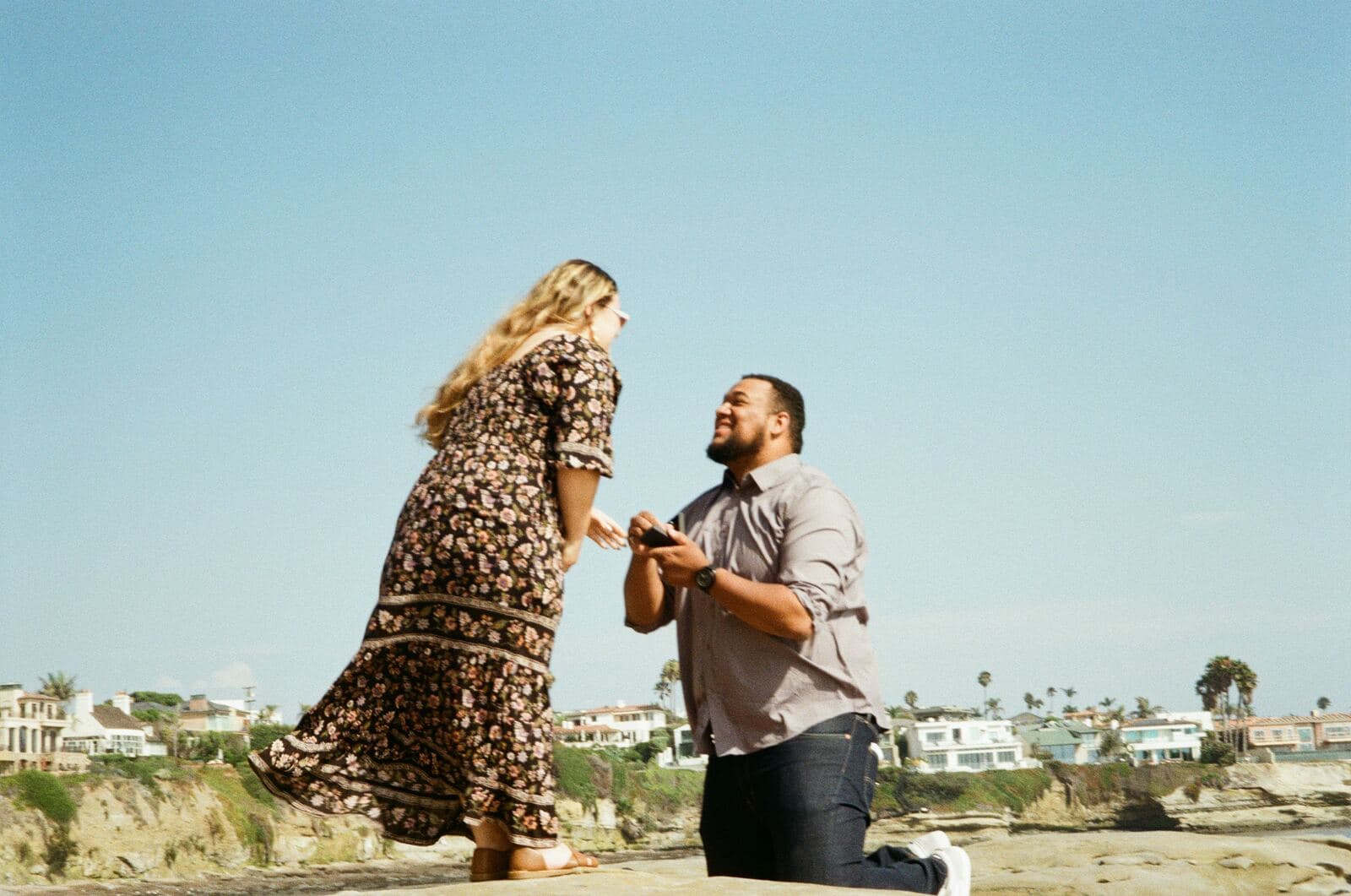man on one knee opening a ring box before a woman in a flowing floral dress on seaside cliffs