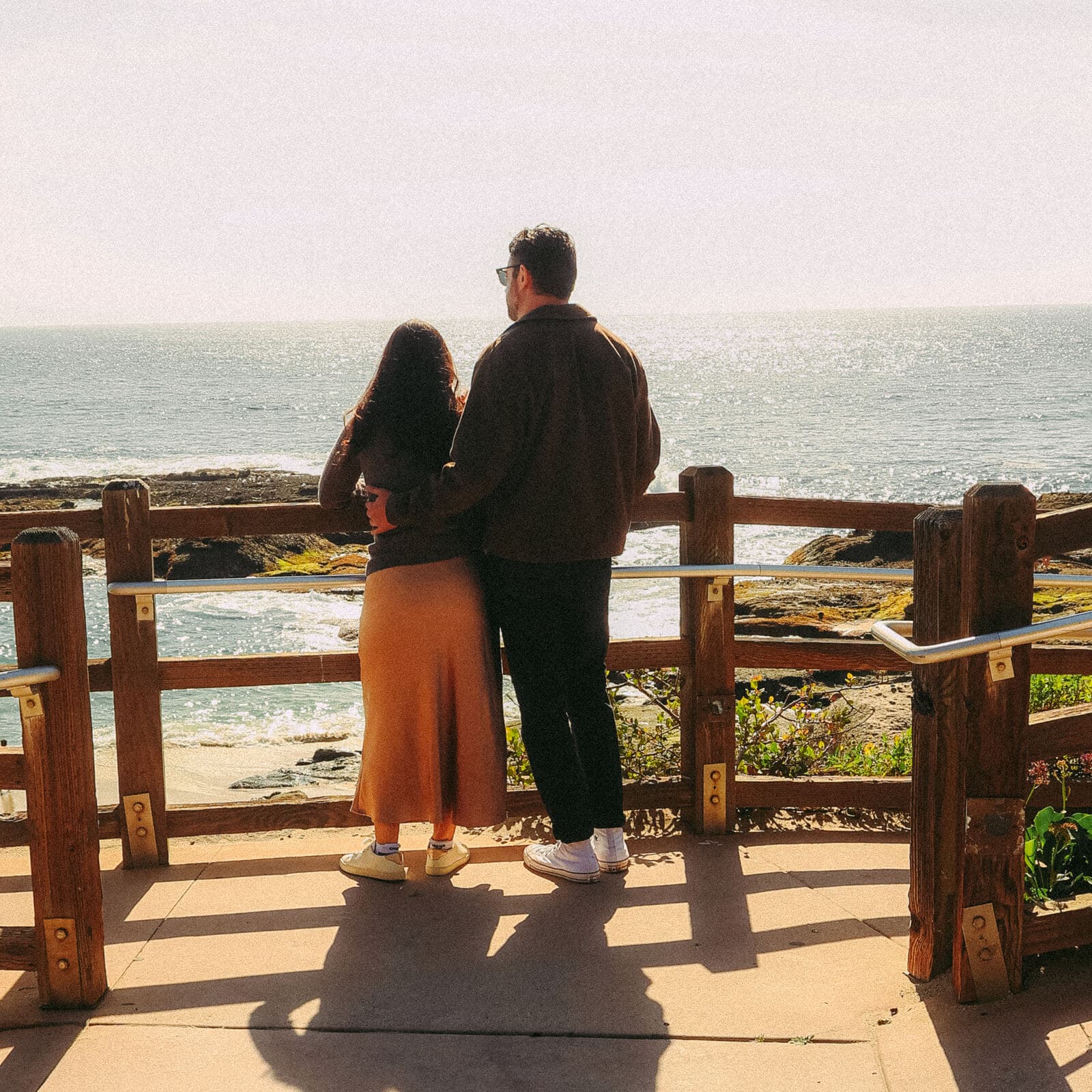 couple standing arm in arm at a wooden railing looking out over the ocean