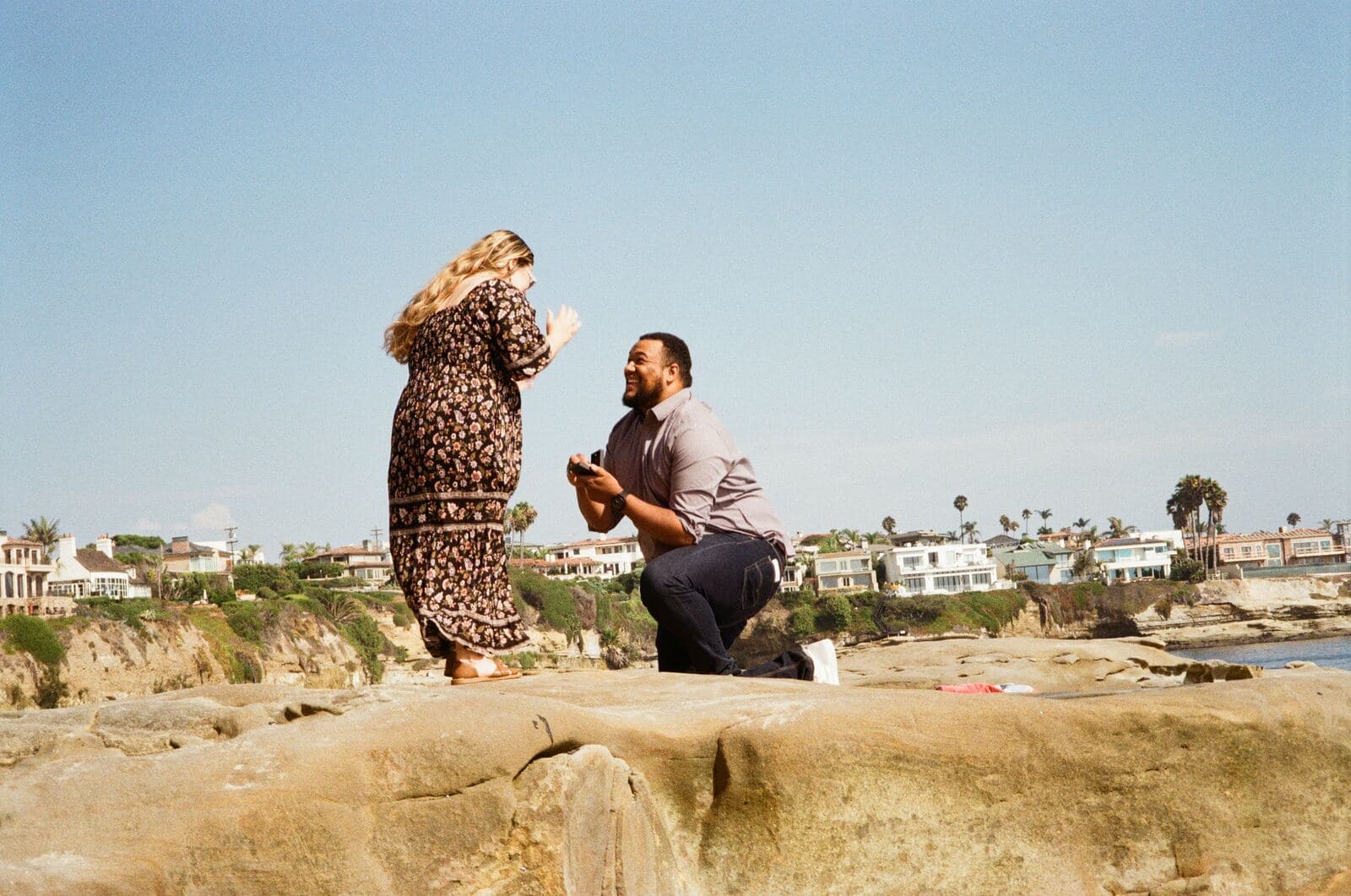 man kneeling with a ring box before a surprised woman on sandstone rocks above a coastal town