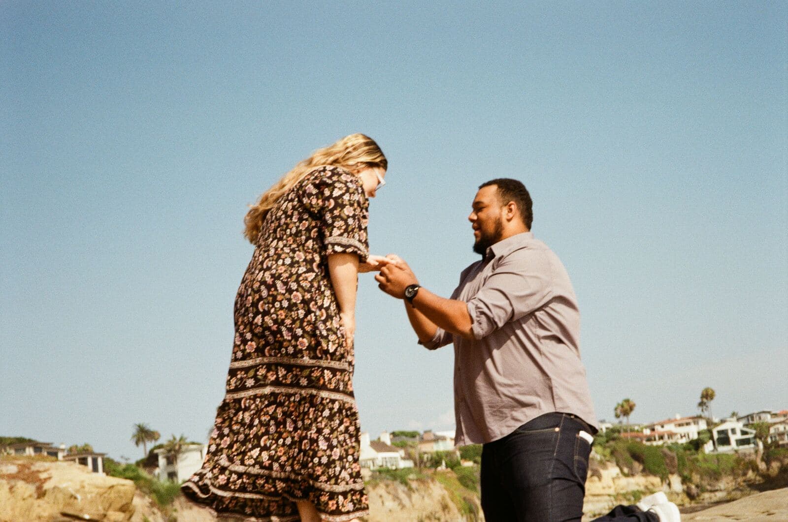 kneeling man placing a ring on a woman's hand on a coastal bluff under a clear sky
