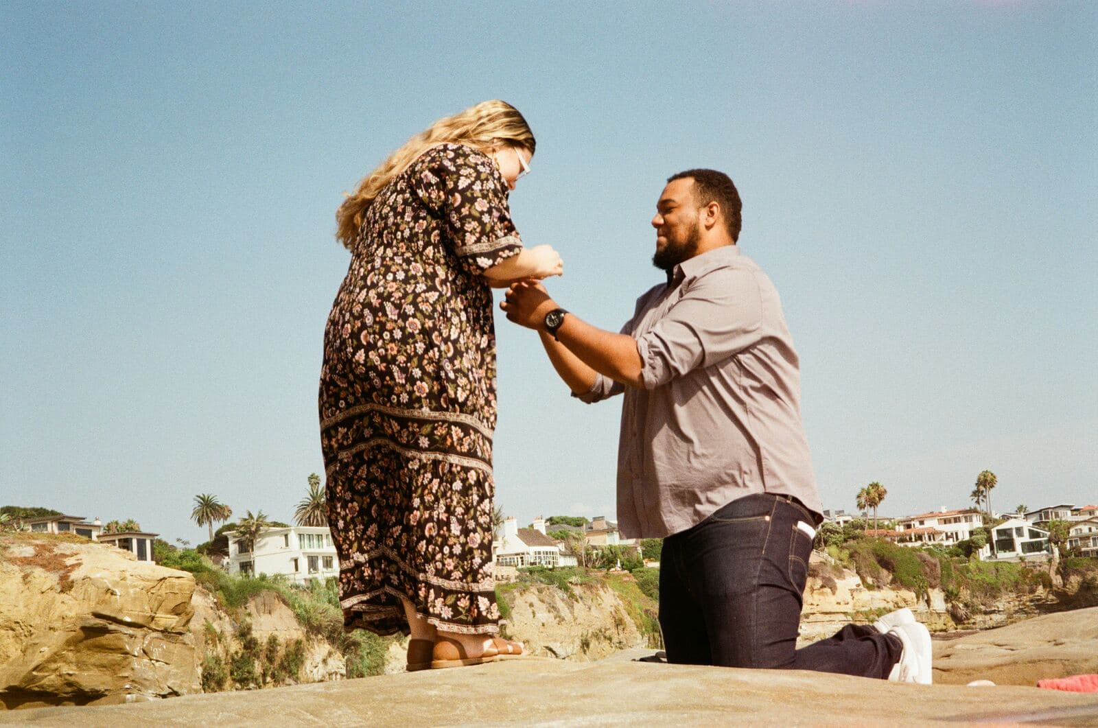 man on one knee holding a woman's hand on coastal rocks with a hillside town behind them