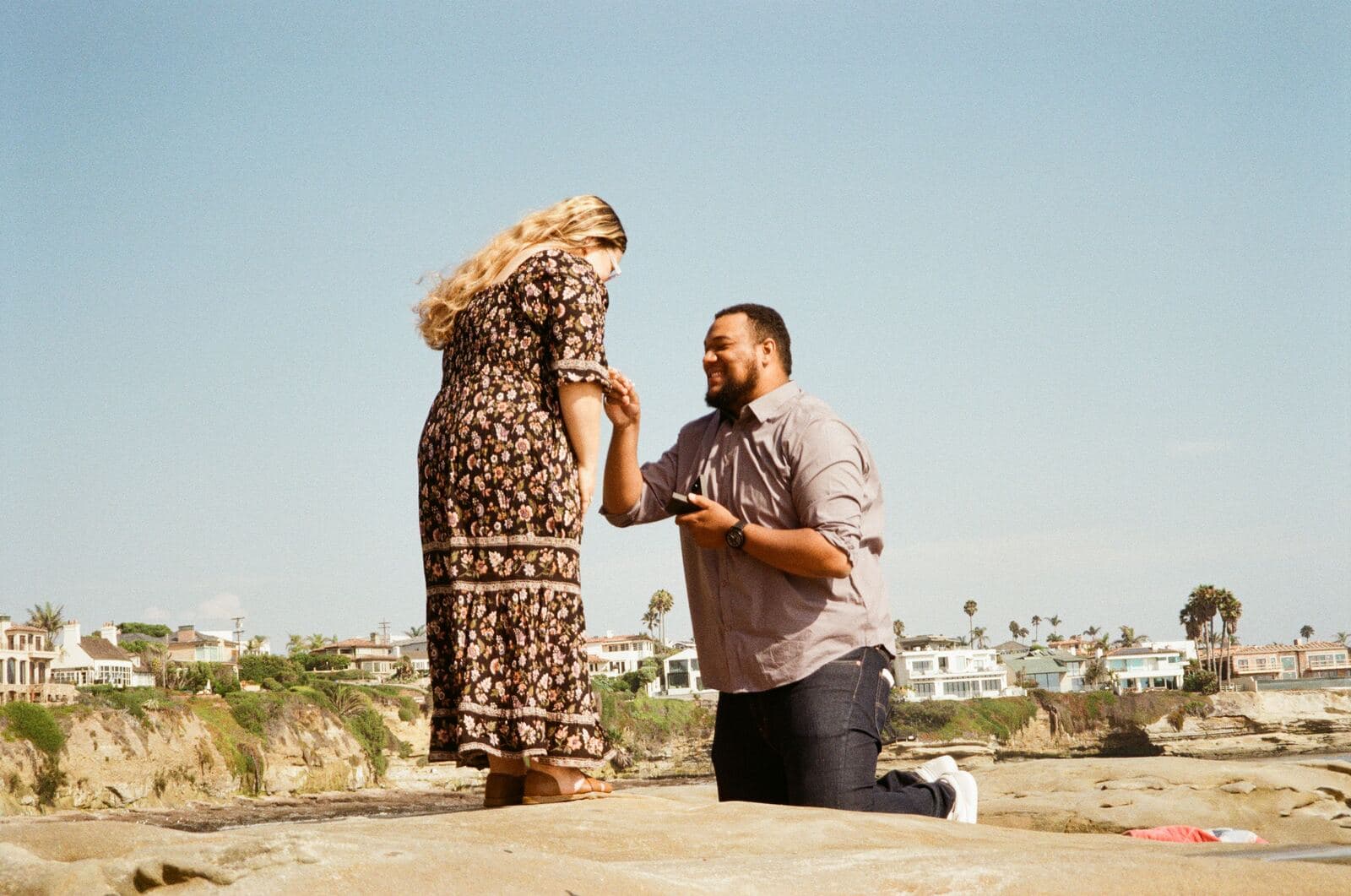man on one knee holding a ring box and the hand of a woman in a floral dress on coastal rocks
