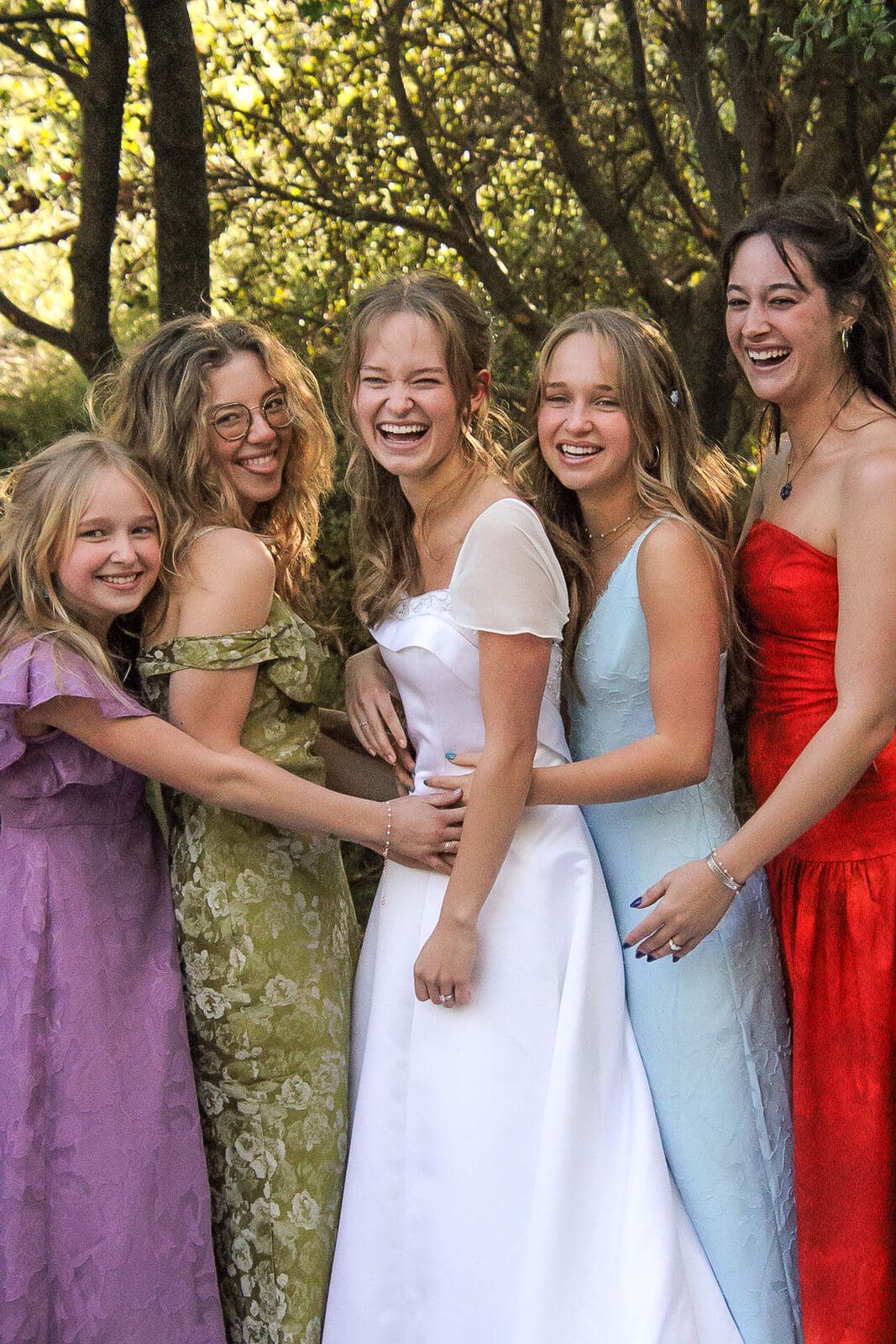 bride laughing in a group hug with four bridesmaids in a sunlit wooded clearing