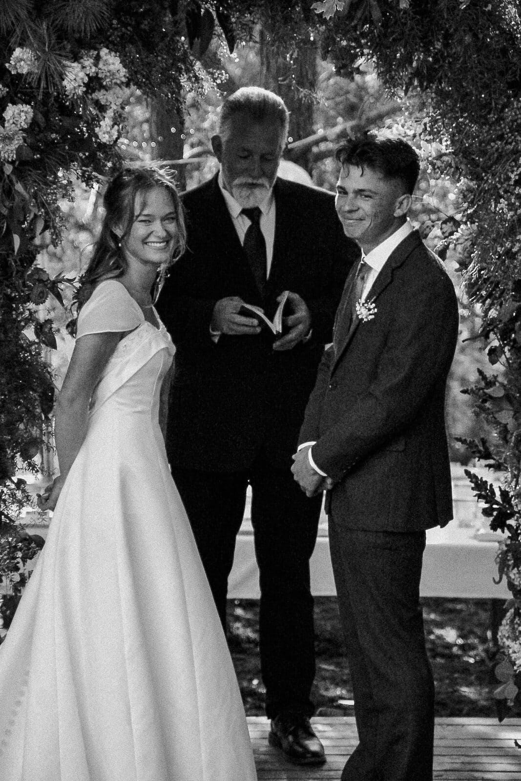 black and white photo of a laughing bride and smiling groom standing with the officiant under a garden arch