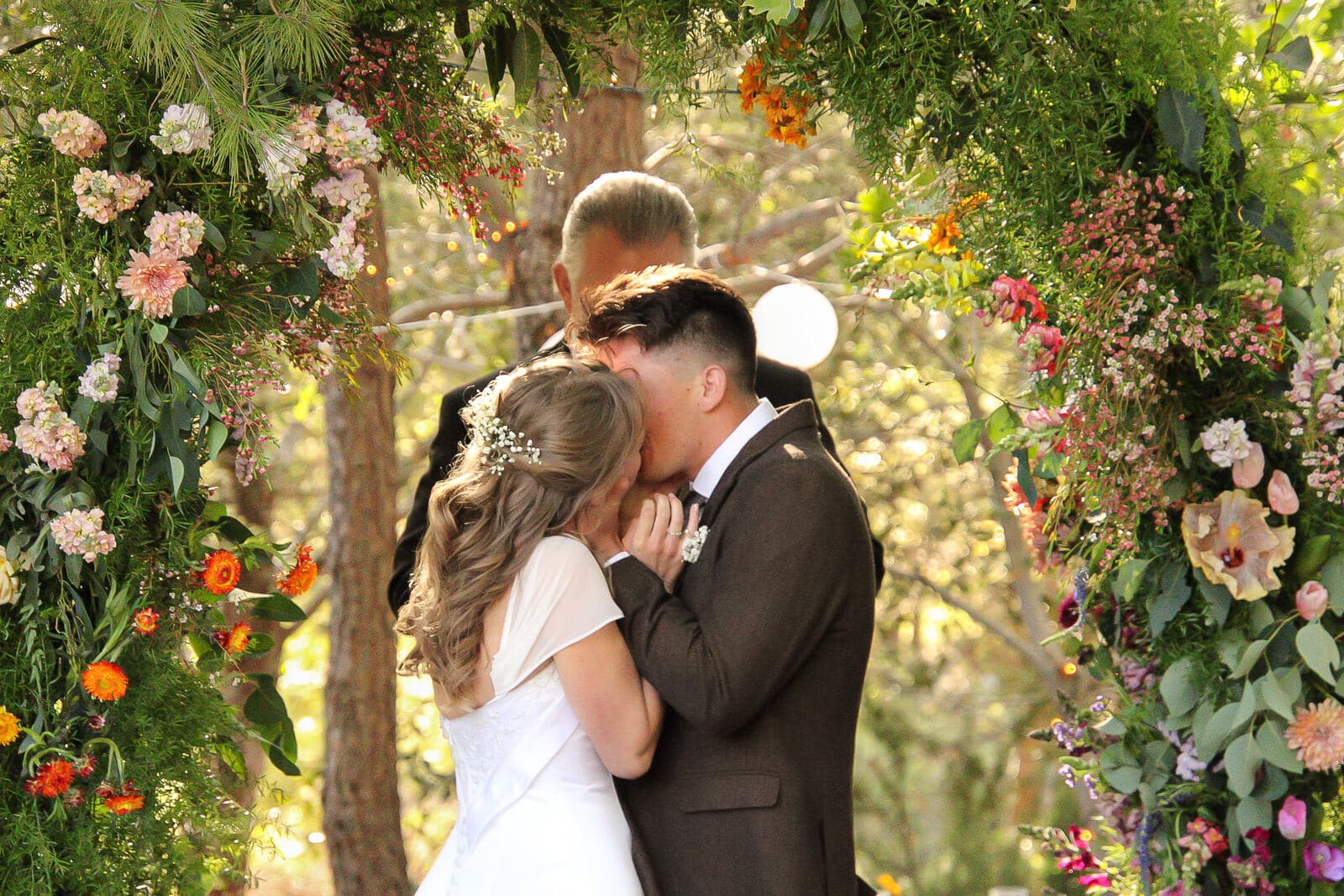 bride and groom sharing their first kiss framed by a colorful floral ceremony arch in the woods