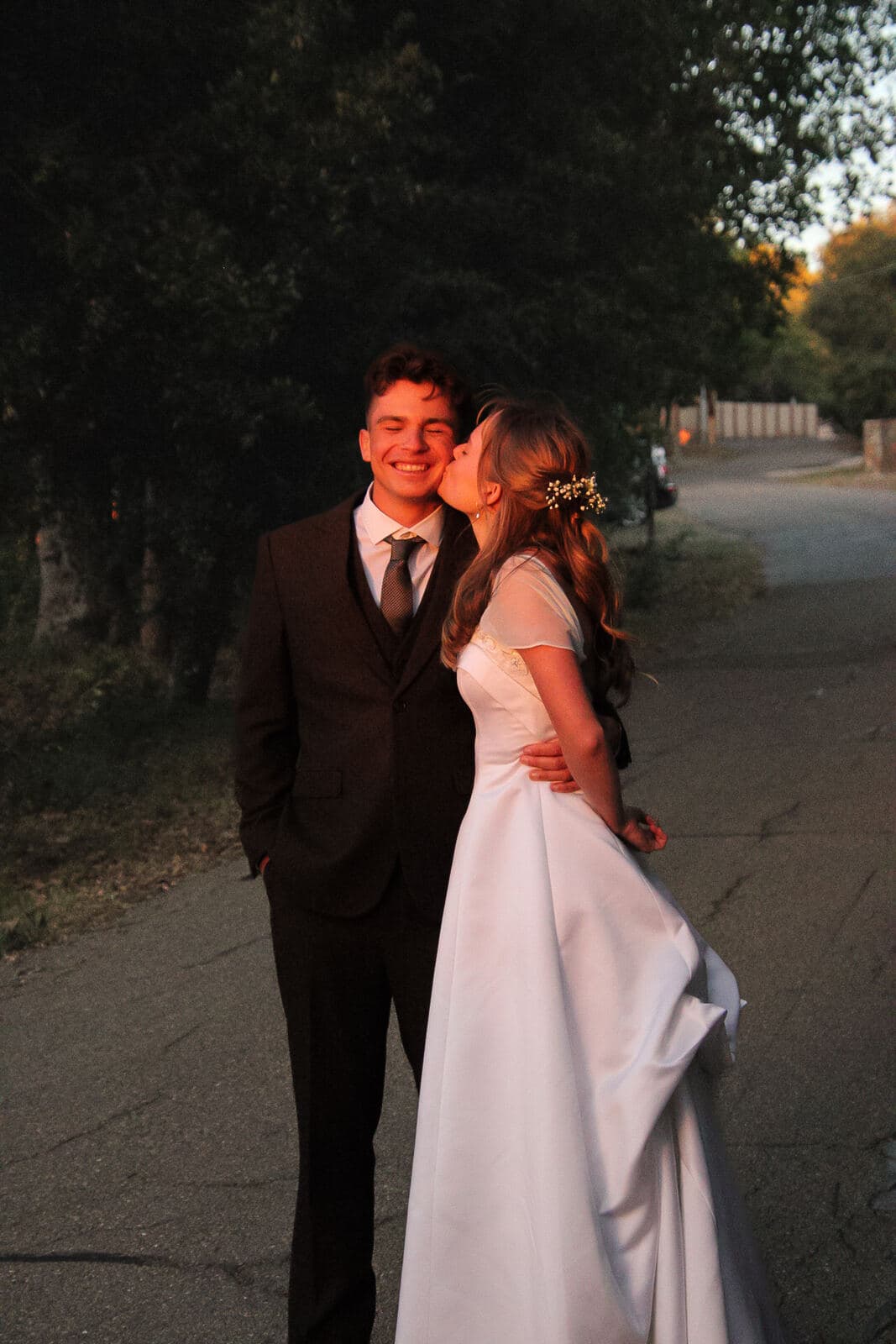 bride kissing groom on the cheek on a tree-lined road at golden hour
